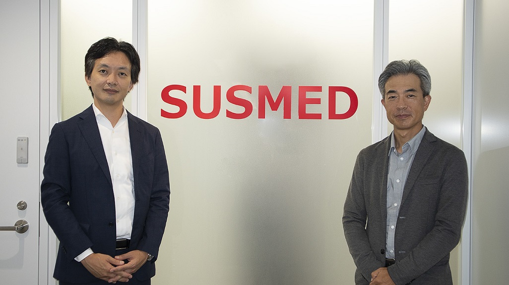 男性二人がSUSMEDと書いてある看板の前で並んで立つ様子