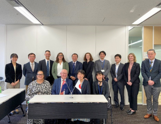 Photo of New Zealand MBIE Deputy Chief Executive Pigou and NEDO Executive Director Iimura smiling in front of the flags of both countries, as well as Maori Trustee Dr. Severn, NEDO, MBIE, MFAT, and GNS Science staff who participated in the exchange of views.
